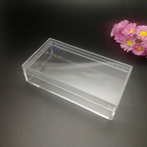 Puede incluir: Una caja rectangular de plástico transparente con tapa. La caja está vacía y se encuentra sobre una superficie negra.