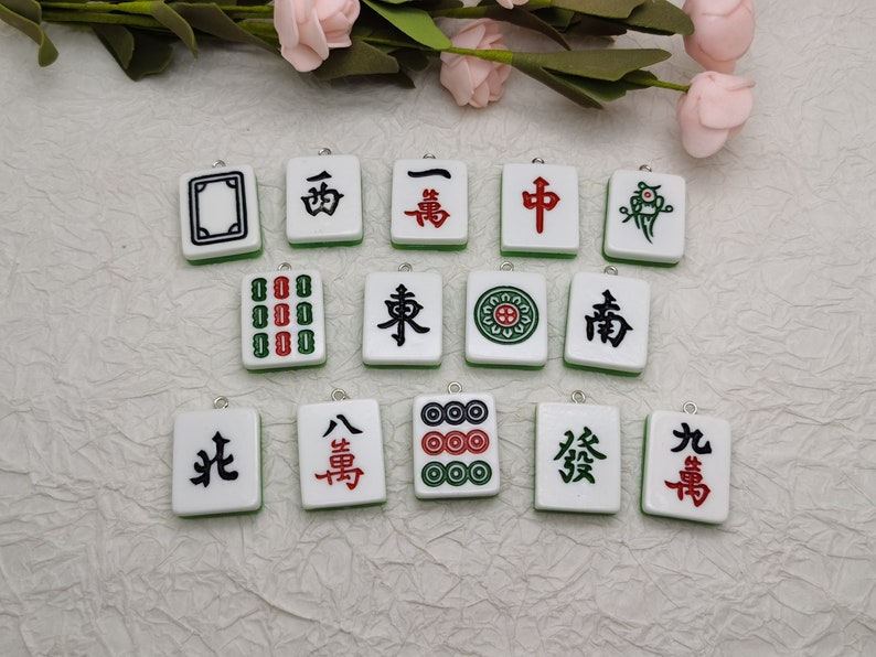 Puede incluir: Un conjunto de 14 amuletos de fichas de mahjong blancas y verdes con caracteres y s&iacute;mbolos chinos negros y rojos. Los amuletos est&aacute;n dispuestos en tres filas sobre una superficie blanca.