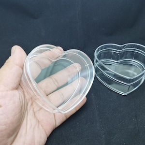 Puede incluir: Dos cajas transparentes de plástico en forma de corazón. La caja de la izquierda está siendo sostenida por una mano. La caja de la derecha está sentada sobre una superficie negra.