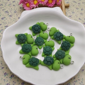 Puede incluir: Un plato blanco con 12 dijes de tortuga verde. Los dijes tienen un caparazón verde y una cara blanca con ojos negros. Cada dije tiene un lazo plateado para sujetarlo a un collar o pulsera.