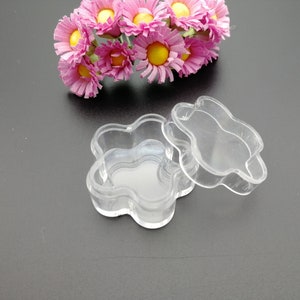 Op de afbeelding: Een transparante, bloemvormige doos van plastic met een deksel. De doos is leeg en heeft een glad oppervlak.