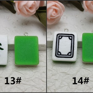 Puede incluir: Dos fichas de mahjong chinas verdes y blancas con dijes plateados. La primera ficha tiene un fondo blanco con caracteres chinos negros. La segunda ficha tiene un fondo blanco con un borde negro y un dise&ntilde;o negro en el interior.