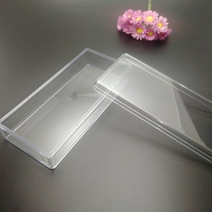 Puede incluir: Una caja rectangular de plástico transparente con tapa. La caja está vacía y la tapa está abierta.