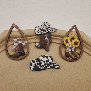 May include: Four wooden cutouts with a cowboy theme. Two of the cutouts are shaped like teardrops and feature a cowboy boot with a cowboy hat. One cutout is a cowboy hat with a black and white cow print design. The other cutout is a pair of cowboy boots with sunflowers.