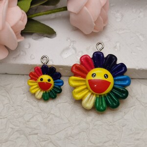 Puede incluir: Dos abalorios de flores coloridos con una cara sonriente. El abalorio más grande tiene un diámetro de unos 5 cm y el más pequeño de unos 2,5 cm. Ambos abalorios tienen un lazo plateado para sujetarlos a un collar o una pulsera.