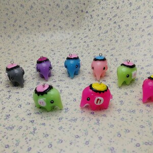 Könnte beinhalten: Acht kleine Elefantenfiguren aus buntem Polymer Clay. Jeder Elefant hat eine andere Farbe und einen schwarz-weißen Hut mit einer kleinen Schlaufe oben.
