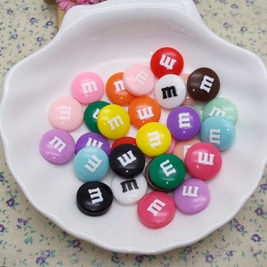 Könnte beinhalten: Eine Sammlung bunter, plastik-bonbonförmiger Knöpfe mit dem Buchstaben "M" darauf. Die Knöpfe sind in verschiedenen Farben, darunter Rot, Orange, Gelb, Grün, Blau, Lila, Rosa und Braun.