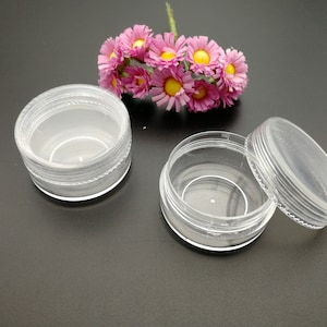 Op de afbeelding: Twee transparante plastic containers met deksels. De containers zijn rond en hebben een gladde, glanzende afwerking. De deksels zijn ook transparant en hebben een licht getextureerde afwerking.