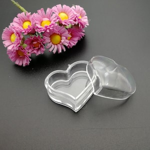 Op de afbeelding: Een doorzichtige, hartvormige plastic doos met een deksel, gedeeltelijk open. De doos staat op een donkere ondergrond, met een boeket roze en gele bloemen op de achtergrond. De doos is waarschijnlijk voor kleine items.