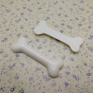 Pode incluir: Dois brinquedos de plástico branco em forma de osso de cachorro. Os ossos são de tamanhos diferentes.