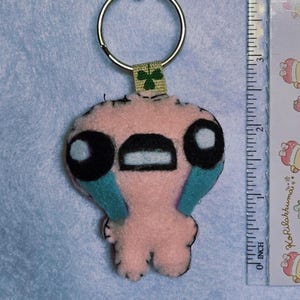 Può includere: Portachiavi fatto a mano in feltro rosa con un personaggio dei cartoni animati con grandi occhi neri, bocca nera e dettagli turchese. Il portachiavi include un anello di metallo e una piccola etichetta verde a forma di trifoglio. Un righello è accanto al portachiavi.
