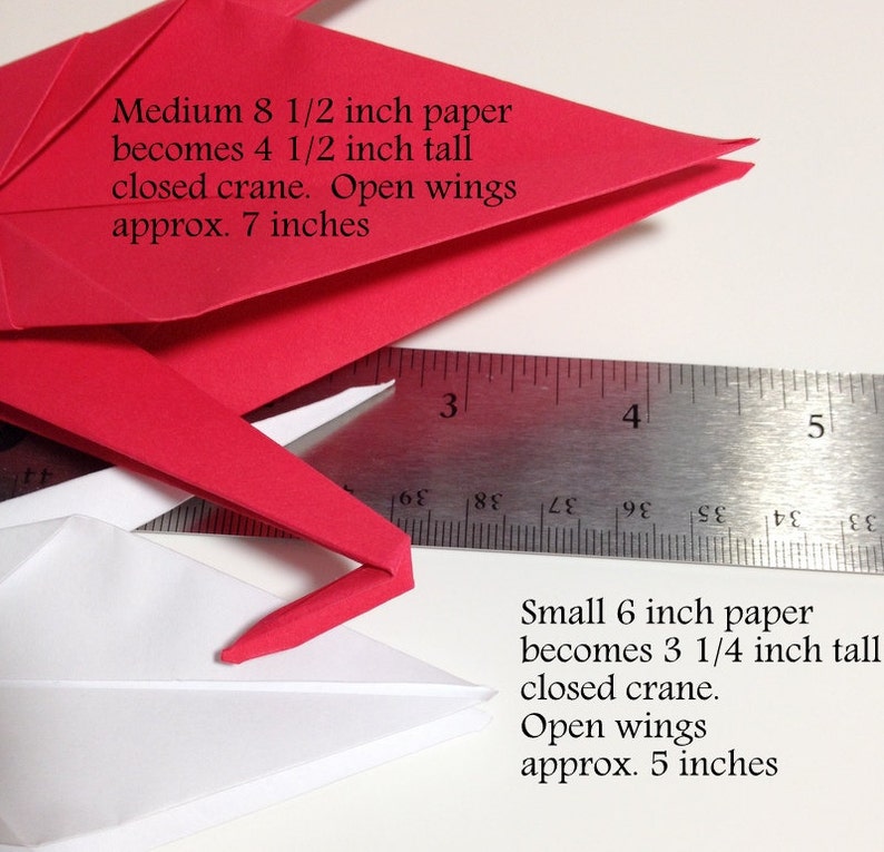 May include: Close-up of red and white origami cranes with a ruler for scale. The text indicates the size of paper used and the dimensions of the finished cranes. The red crane is made from 8 1/2 inch paper and the white crane from 6 inch paper.