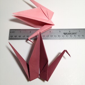 May include: Two origami cranes, one pink and one burgundy, are displayed with a ruler. The pink crane is folded in a closed position, while the burgundy crane is open. The ruler shows measurements in inches.