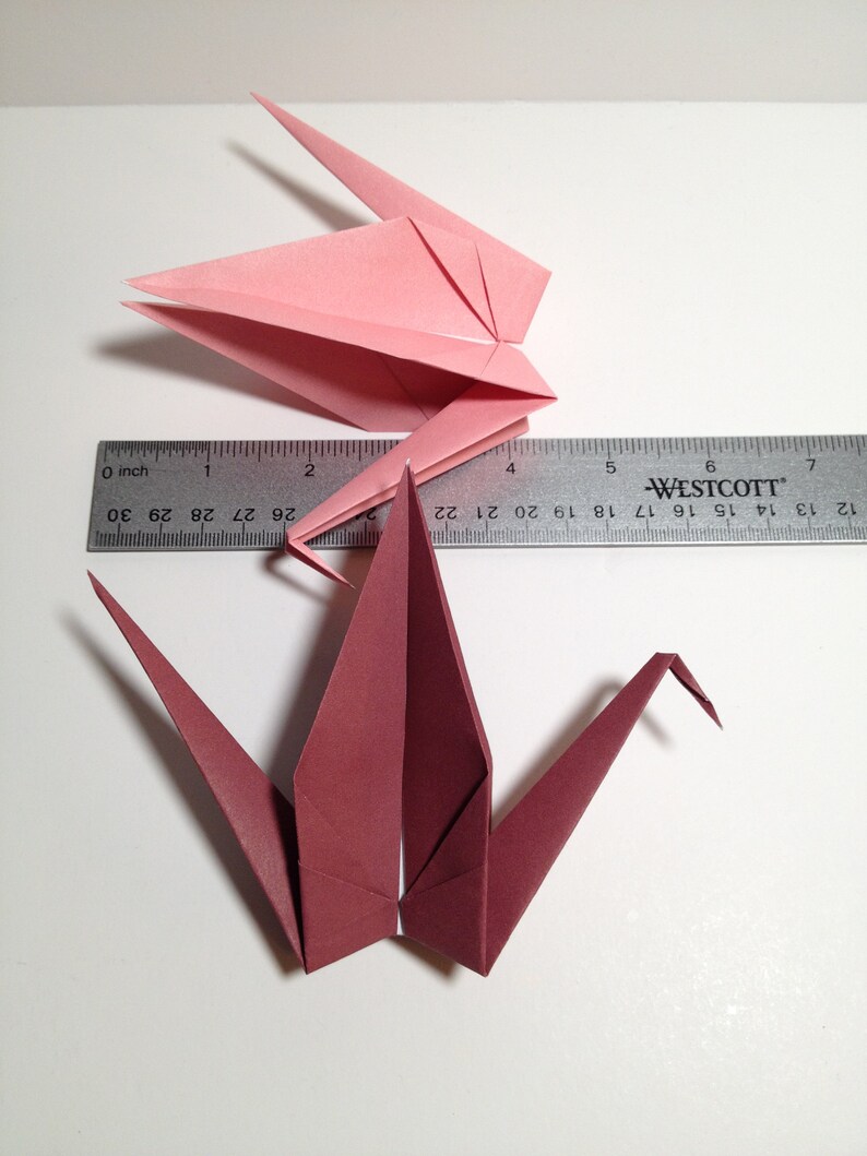 May include: Two origami cranes, one pink and one burgundy, are displayed with a ruler. The pink crane is folded in a closed position, while the burgundy crane is open. The ruler shows measurements in inches.
