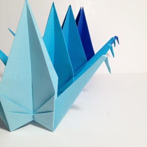 May include: A light blue origami crane with a series of triangular peaks in varying shades of blue. The paper craft is set against a white background, showcasing the detailed folds and geometric shapes of the artwork.