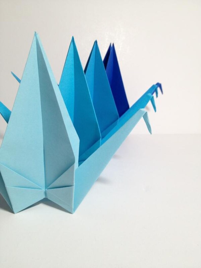 May include: A light blue origami crane with a series of triangular peaks in varying shades of blue. The paper craft is set against a white background, showcasing the detailed folds and geometric shapes of the artwork.