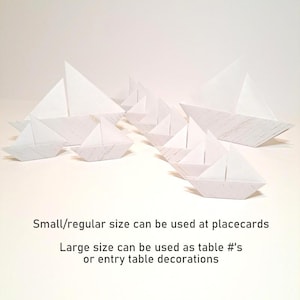 May include: A collection of white paper origami boats of varying sizes. The boats are arranged on a white surface. Text on the image states that the small boats can be used as placecards and the large boats as table numbers or decorations.