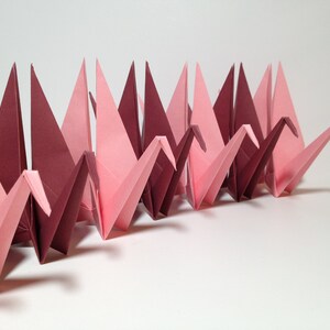 May include: A row of origami cranes in varying shades of pink and burgundy. The paper birds are folded in a repeating pattern, creating a visually appealing display. The cranes are arranged on a white surface.
