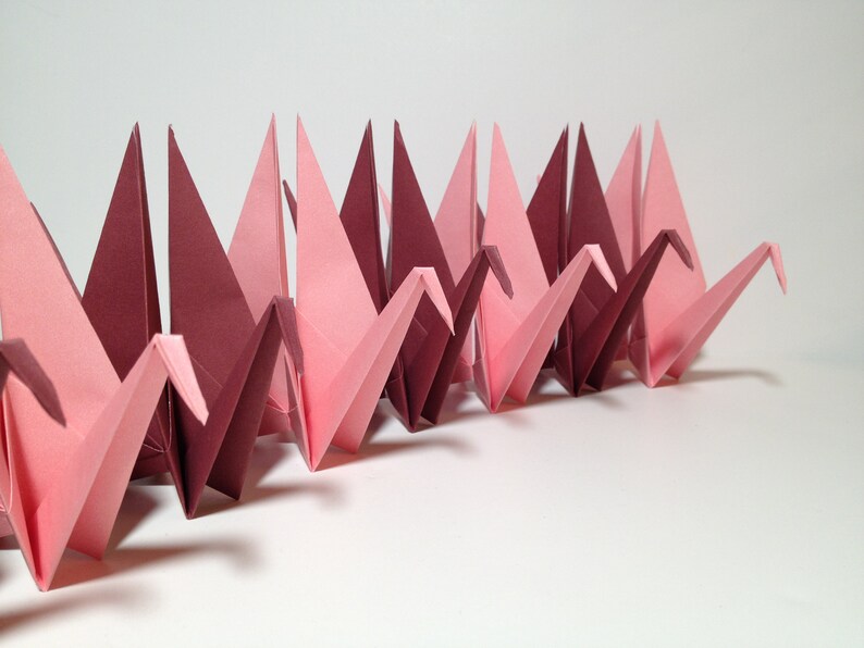 May include: A row of origami cranes in varying shades of pink and burgundy. The paper birds are folded in a repeating pattern, creating a visually appealing display. The cranes are arranged on a white surface.