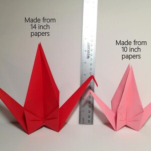 May include: Two origami cranes, one red and one pink, are displayed next to a ruler. The red crane is made from 14-inch paper, and the pink crane is made from 10-inch paper. The text on the image indicates the paper size used.