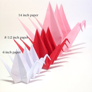 May include: Origami paper cranes in a gradient of colors, from white to pink and red. The cranes are made from 6-inch, 8.5-inch, and 14-inch paper. The cranes are arranged in a row, showcasing the size difference.