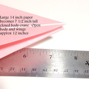 May include: A folded pink paper crane with a ruler. The text on the image says: Large 14 inch paper becomes 7 1/2 inch tall closed body crane. Open body and wings approx 12 inches.