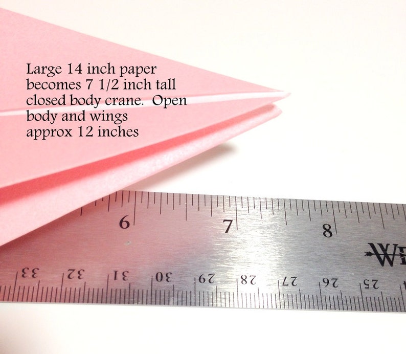 May include: A folded pink paper crane with a ruler. The text on the image says: Large 14 inch paper becomes 7 1/2 inch tall closed body crane. Open body and wings approx 12 inches.