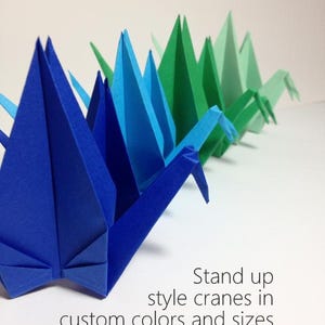 May include: A row of origami cranes in shades of blue and green. The paper cranes are standing upright, showcasing their folded wings and pointed beaks. Text on the image reads "Stand up style cranes in custom colors and sizes."
