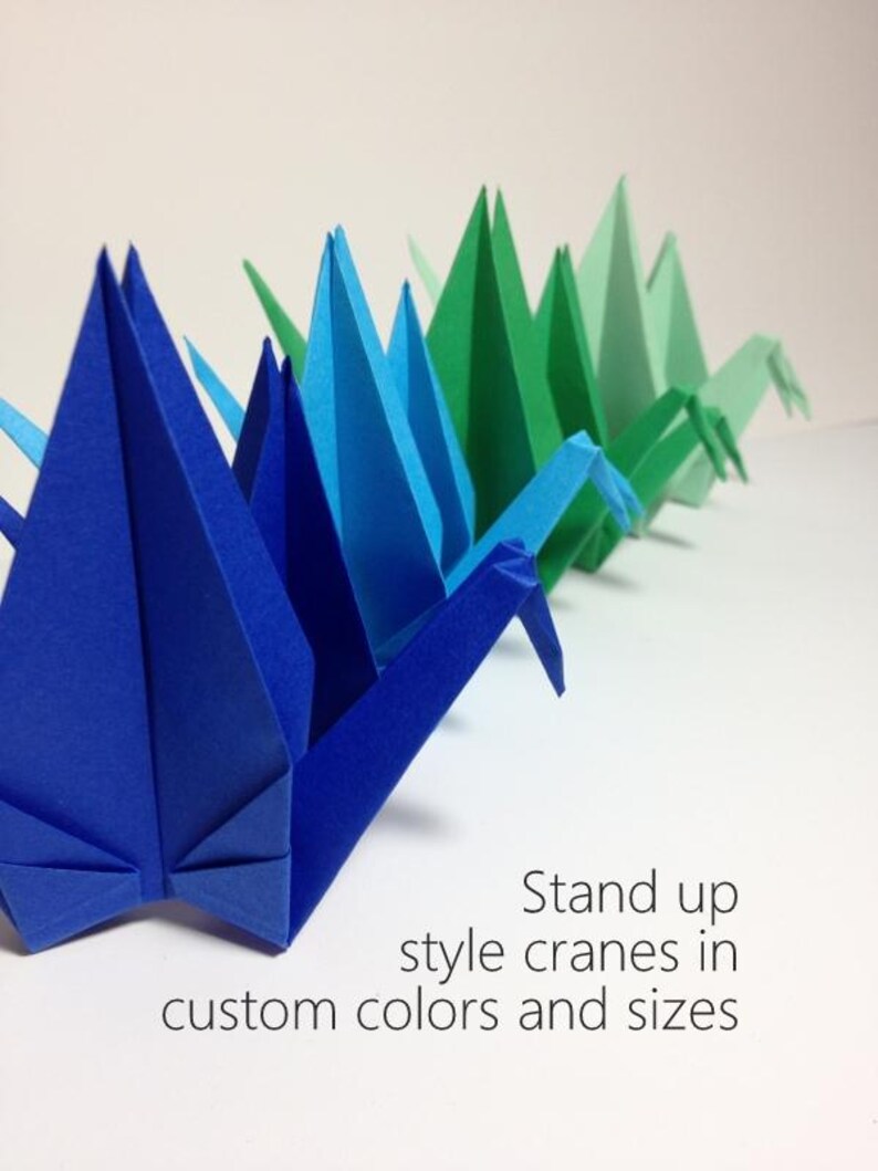 May include: A row of origami cranes in shades of blue and green. The paper cranes are standing upright, showcasing their folded wings and pointed beaks. Text on the image reads "Stand up style cranes in custom colors and sizes."