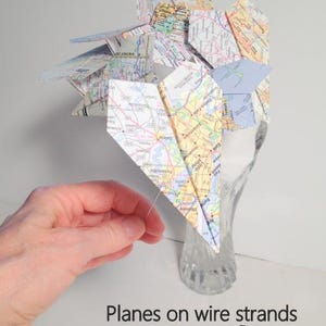 May include: A collection of paper airplanes folded from colorful road maps, each attached to a thin wire strand. The text Planes on wire strands as inserts into flower centerpieces is printed at the bottom of the image.