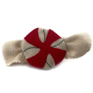 May include: A red and white felt candy with a white felt wrapper. The candy is shaped like a peppermint and has a red center with white stripes.
