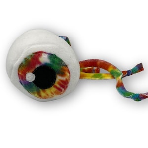 May include: A plush toy shaped like a large eye with a tie-dye pattern. The eye is white with a black pupil and a rainbow tie-dye pattern around the pupil. The toy has a long, thin, rainbow tie-dye tail.