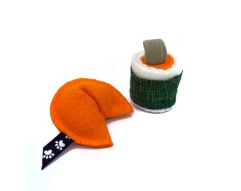 May include: Two plush dog toys shaped like a fortune cookie and a sushi roll. The fortune cookie is orange with a black and white paw print ribbon. The sushi roll is green, white, and orange.