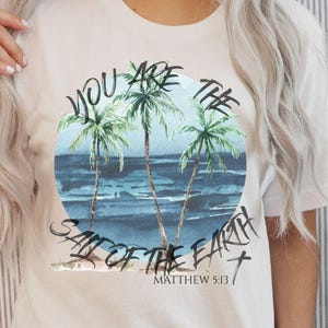 May include: Cream-colored t-shirt featuring a watercolor design of palm trees on a beach with the text "YOU ARE THE SALT OF THE EARTH" and "MATTHEW 5:13". The design is centered on the shirt.