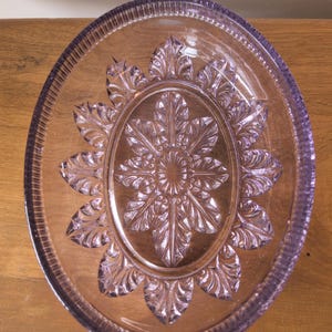 May include: A clear, oval glass dish with a lavender hue. The dish has a detailed floral pattern in the centre and a textured rim. The design is symmetrical and the dish is on a wooden surface.