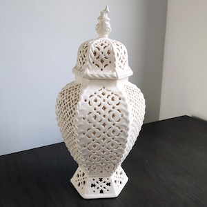 May include: A white ceramic decorative jar with a lid. The jar features intricate cut-out patterns and a textured surface. The lid is topped with a decorative finial. The jar has a hexagonal base.