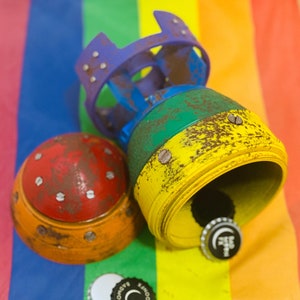 May include: A set of three colorful, round, metal bottle stoppers with a rainbow background. The stoppers have a rusty, aged look and are decorated with small metal dots. One stopper has a blue, green, and yellow color scheme, another is red and orange, and the third is a solid red color. The stoppers are on a rainbow-colored background.