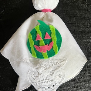May include: A handmade Halloween decoration featuring a white fabric ghost with a watermelon-themed face. The ghost has a round head, a pink ribbon, and a green and pink felt watermelon design. A white lace handkerchief is attached.