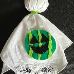 May include: A handmade Halloween decoration. It features a white ghost-like figure with a green and black watermelon-themed face. The ghost has a round head and a flowing body, with a green ribbon around the neck. The bottom edge has a decorative cut-out.