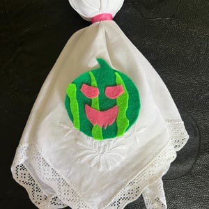 May include: A handmade ghost doll with a white fabric body and head, secured with a pink ribbon. The doll's body features a green and pink watermelon applique, and the bottom edge is trimmed with white lace.