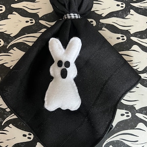 May include: A black cloth napkin with a white felt ghost decoration. The napkin is folded and tied with a black and white gingham ribbon. The background features a pattern of white ghost figures on a black surface.