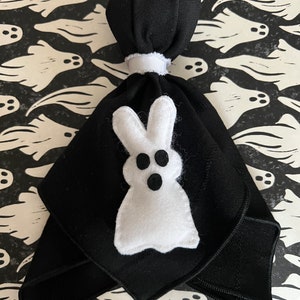 May include: A black fabric ghost with a white felt ghost bunny face. The ghost is tied at the top with a white ribbon. The background is a black and white pattern of ghosts.