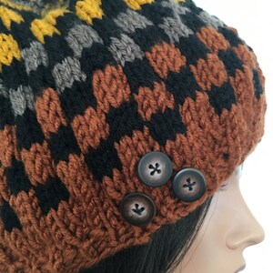 May include: A brown, black, gray, and yellow knitted beanie with three brown buttons. The beanie is made with a chunky yarn and has a textured pattern.