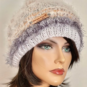 May include: White, gray, and beige knitted hat with a fluffy texture and a brown leather tag that says "Hand Knits & Love" with a small paw print.
