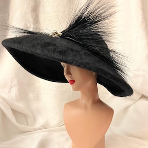 May include: A wide-brimmed black hat with a textured surface, featuring a cluster of long, dark, wispy feathers and a small, ornate metallic embellishment with a central pearl-like accent positioned at the top center of the crown.
