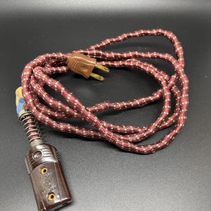 May include: A vintage brown and white striped electrical cord with a two-prong plug on one end and a three-prong plug on the other end. The cord has a spring on the three-prong plug end.
