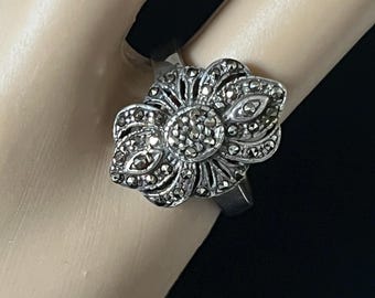 May include: A silver-toned ring featuring a detailed flower design. The ring's petals are adorned with small, dark stones, creating a textured and eye-catching floral pattern. The ring is shown on a hand.