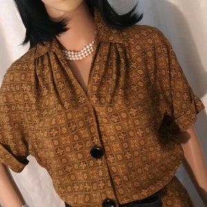 May include: A vintage-style two-piece outfit in shades of brown, featuring a geometric floral pattern. The short-sleeved top has large black buttons and a collared neckline. A black belt cinches the waist. The mannequin is also wearing a pearl necklace and bracelet.