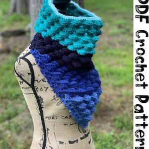 May include: A crocheted cowl in shades of blue and teal. The cowl is made with a textured stitch that creates a bumpy effect. The cowl is displayed on a fabric-covered mannequin.