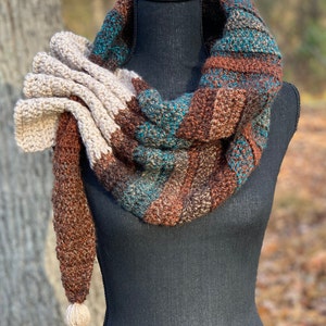 May include: A brown, teal, and cream striped crocheted scarf with a large cream crocheted flower accent and a long cream tassel.